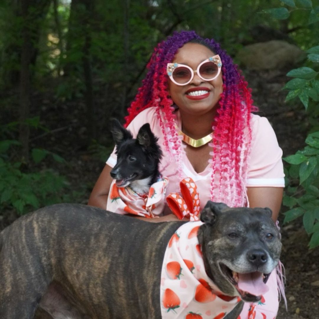 Bre Hart, Owner of You Can Leave Pet Sitting Services A woman with vibrant hair poses outdoors with two dogs dressed in matching scarves.